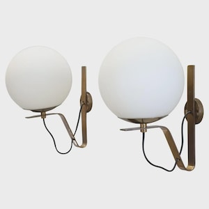 Puede incluir: Dos apliques de pared con pantallas esféricas de vidrio blanco. Los apliques tienen un marco de metal color latón y un cable eléctrico negro. El diseño es minimalista y moderno.