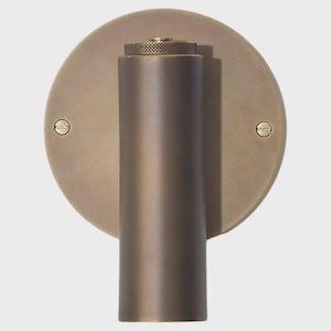 May include: A bronze-colored wall sconce with a circular backplate and a cylindrical arm. The sconce has a textured top and two small screws securing the backplate. The design is minimalist and modern.