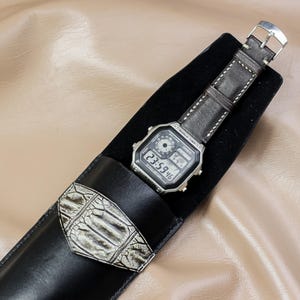 May include: A digital watch with a black face and brown leather strap rests in a black leather case. The watch displays the time 23:59:46. The case has a decorative white and gray alligator skin accent.