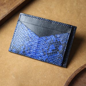 May include: A blue and black leather card holder with a snakeskin pattern. The card holder has a rectangular shape with a black leather pocket and blue snakeskin design. The edges are stitched with black thread.