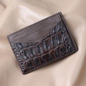 Puede incluir: Un tarjetero de cuero marrón con textura de piel de cocodrilo en la mitad inferior. La mitad superior es de cuero liso, marrón más oscuro. El tarjetero es rectangular y tiene un borde cosido.