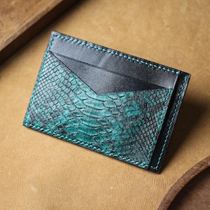 May include: A teal and black leather card holder with a snakeskin pattern. The card holder has a teal stitched border and a black leather pocket. The card holder is rectangular and has a unique design.