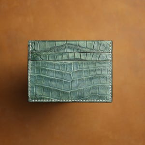 May include: A teal green cardholder with a crocodile skin texture. The rectangular wallet has white stitching around the edges and a slot on the top. The background is a solid brown.