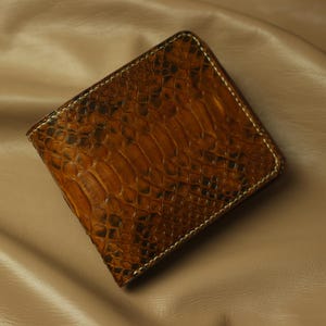 Może przedstawiać: Brązowy portfel dwukrotnie składany ze wzorem skóry węża. Portfel jest wykonany ze skóry i ma teksturowaną powierzchnię. Krawędzie są obszyte białą nicią.