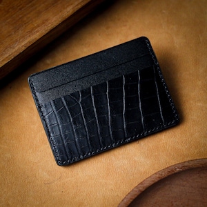 Puede incluir: Tarjetero de cuero negro con textura de cocodrilo en la parte delantera. El tarjetero tiene múltiples ranuras y es rectangular. Los bordes están cosidos con hilo negro. El fondo es una superficie de cuero marrón claro.