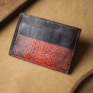 Op de afbeelding: Een handgemaakte kaarthouder met een opvallend ontwerp. Het bovenste gedeelte is zwart leer, terwijl de onderste helft een roodbruin slangenpatroon heeft. De randen zijn zorgvuldig gestikt met bruin garen.