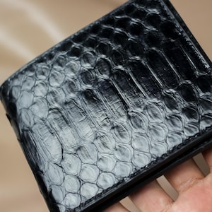 Op de afbeelding: Een zwarte bi-fold portemonnee met een textuur, schubachtig patroon. De portemonnee lijkt van leer te zijn, met een subtiele glans. Het ontwerp suggereert een reptielenhuid, wat een unieke textuur aan het accessoire toevoegt.