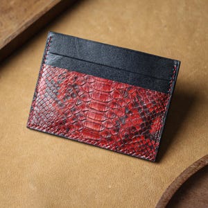 May include: A rectangular card holder featuring a red snakeskin pattern and black leather detailing. The card holder is stitched with red thread, showcasing a minimalist design. The product is likely made of genuine leather.