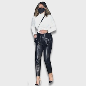 May include: A white long-sleeved mock neck top paired with high-waisted black leather trousers featuring a tie-waist belt. The outfit includes a black face mask, a thin crossbody bag strap, and black pointed-toe high-heeled shoes.