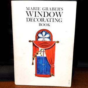 May include: A white book with the title "Marie Graber's Window Decorating Book" in black text. The cover features an illustration of a red and white window with a blue shutter and a stained glass design.