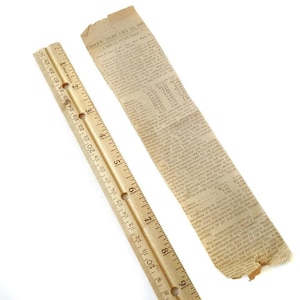 May include: A wooden ruler with inch markings and a piece of aged newspaper. The ruler is light brown with black markings and holes for hanging. The newspaper fragment has text and is torn at the edges.