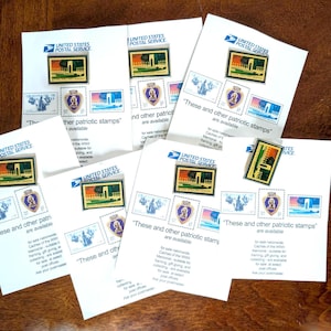 May include: Several white envelopes with patriotic stamps from the United States Postal Service are displayed on a wooden surface. The stamps feature images of monuments and the Purple Heart. Text on the envelopes reads "These and other patriotic stamps are available."