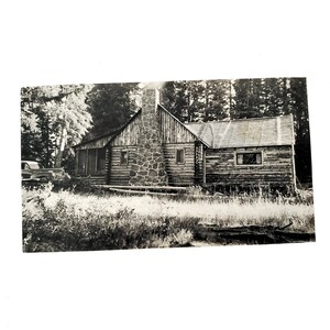 May include: A black and white photo of a rustic log cabin with a stone chimney. The cabin has a wooden roof and windows. There are trees in the background.