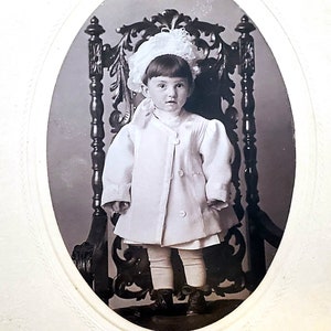 May include: A black and white portrait of a young child wearing a white dress and a white bonnet, sitting in a carved wooden chair.