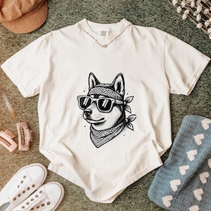 Op de afbeelding: Een crèmekleurig T-shirt met korte mouwen en een zwart-witte grafische illustratie van een hond met een zonnebril en een bandana met patroon om de nek. Het ontwerp is gecentreerd op de borst van het T-shirt met een ronde hals.