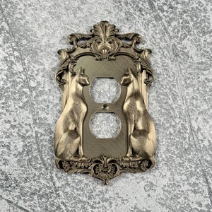 May include: A metallic gold-toned light switch wall plate featuring an ornate, decorative frame with intricate scrollwork and two sculpted cats sitting in profile on either side of the central duplex electrical outlet openings.