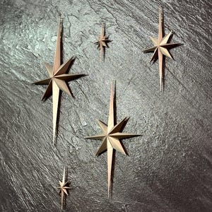 May include: A collection of metallic star-shaped wall decorations in varying sizes. The stars have a bronze and gold color scheme, with long, pointed rays extending from the central star shape. The stars are arranged on a dark, textured surface.