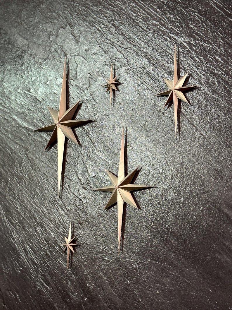 May include: A collection of metallic star-shaped wall decorations in varying sizes. The stars have a bronze and gold color scheme, with long, pointed rays extending from the central star shape. The stars are arranged on a dark, textured surface.