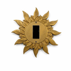 May include: A decorative light switch cover in the shape of a golden sunflower. The sunflower has detailed petal edges and a circular center with a black switch opening. The cover is mounted on a white wall.