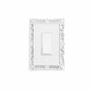 May include: White, ornate light switch cover with a rectangular switch. The cover has a detailed, floral design around the edges. The switch and cover are set against a white background.