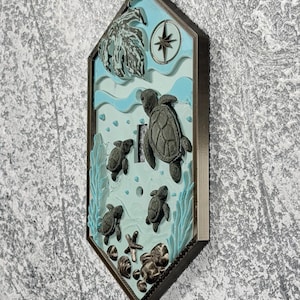 May include: A hexagonal light switch cover featuring a raised, three-dimensional ocean scene with dark grey sea turtles, a starfish, and shells against a light blue background. The design includes a compass rose and seaweed accents in a dark frame.