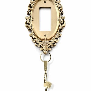 Può includere: Un portachiavi dorato e ornato con una cornice ovale e decorazioni. Una chiave è appesa a un anello nella parte inferiore. Il portachiavi è progettato per essere montato a parete.