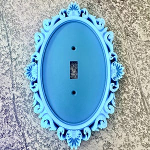 May include: A light blue oval light switch cover plate featuring an ornate, raised scrollwork border. The plate has a central rectangular cutout for a toggle switch and two circular screw holes positioned vertically above and below the center.
