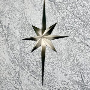 May include: A metallic silver starburst decoration with eight points. The star has a textured surface and is set against a mottled gray background. The star's design is symmetrical, with sharp, elongated points radiating outwards.