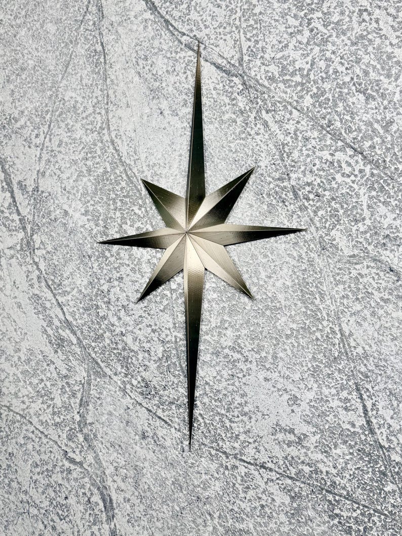May include: A metallic silver starburst decoration with eight points. The star has a textured surface and is set against a mottled gray background. The star's design is symmetrical, with sharp, elongated points radiating outwards.
