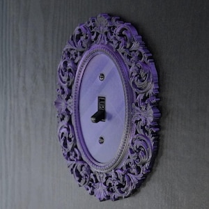 May include: A decorative purple light switch cover with an ornate, oval frame. The frame features intricate floral and scrollwork designs. The switch itself is black, and the cover is mounted on a dark gray wall.