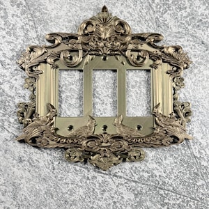 Pode incluir: Uma placa para interruptor triplo com acabamento em dourado metálico, apresentando relevos intrincados de folhagens e quatro pássaros cardeais pousados na borda inferior. A placa retangular possui três aberturas verticais.