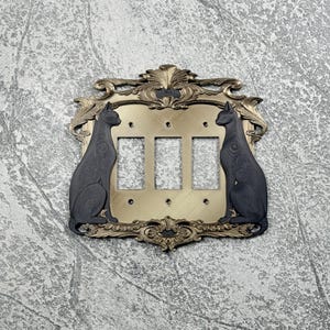 May include: A triple light switch cover plate featuring a metallic gold finish with ornate, scrolling edges and two matte black cat silhouettes facing inward on either side. The rectangular plate includes three vertical cutouts for switches.