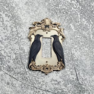 Op de afbeelding: Een rechthoekige lichtschakelaarafdekplaat met een sierlijk goudkleurig frame met ingewikkeld krulwerk en twee matzwarte ravenfiguren die tegenover elkaar staan aan weerszijden van de centrale rechthoekige opening voor de schakelaar.