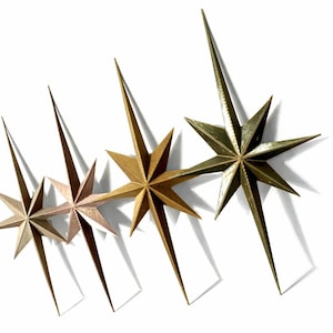 May include: Four metallic starburst wall decor pieces in varying shades of gold and bronze. Each star has multiple points and long, pointed extensions. The stars are arranged diagonally against a white background.