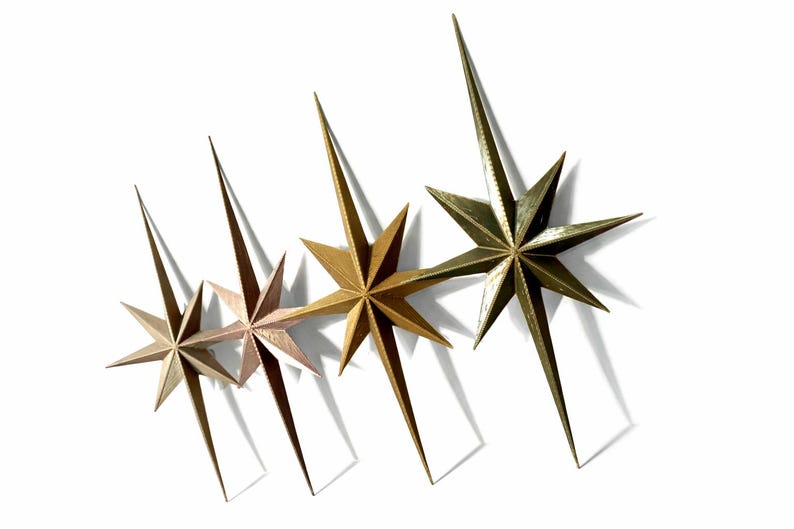 May include: Four metallic starburst wall decor pieces in varying shades of gold and bronze. Each star has multiple points and long, pointed extensions. The stars are arranged diagonally against a white background.