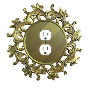 May include: Ornate gold-colored electrical outlet cover with a decorative floral design. The cover features a circular center with two standard electrical outlets and is surrounded by an elaborate, swirling pattern. The overall aesthetic is vintage and elegant.