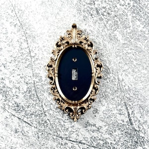 May include: An oval light switch plate featuring an ornate gold-toned frame with intricate scrollwork and leaf-like detailing. The center of the plate is a solid matte black oval insert with a rectangular cutout for a toggle switch and two screw holes.