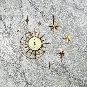 May include: A gold-toned metal light switch plate featuring a celestial design with a crescent moon and sunburst rays, accompanied by seven separate star-shaped wall decorations of varying sizes, all arranged in a cluster on a textured grey surface.