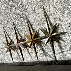 May include: Four metallic starburst wall decor pieces in varying shades of gold and bronze are arranged against a textured, gray background. Each star has eight points and a long, pointed center.