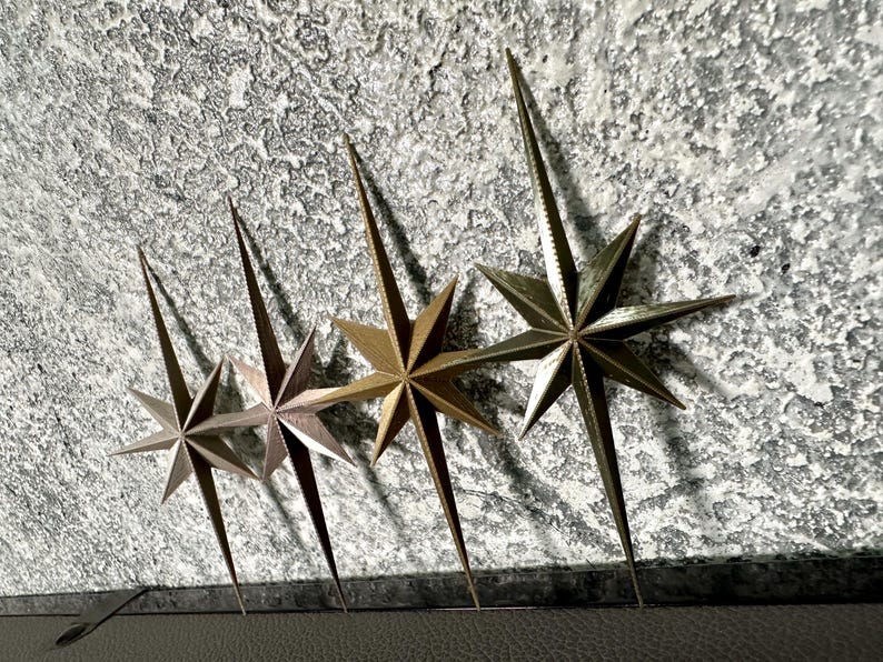 May include: Four metallic starburst wall decor pieces in varying shades of gold and bronze are arranged against a textured, gray background. Each star has eight points and a long, pointed center.