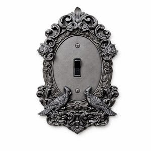 May include: Ornate, pewter-toned light switch cover with an oval center and decorative frame. The frame features floral and scrollwork designs, with two bird figures at the bottom. The switch is black.