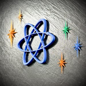 May include: A blue atom symbol and several star-shaped ornaments in shades of orange, green, and blue are arranged on a dark gray surface. The atom symbol is the central focus, with the stars scattered around it.