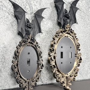 May include: Two oval light switch plates featuring ornate, metallic gold-toned scrollwork frames. Each plate is topped with a three-dimensional black bat figure with outstretched wings. One plate has a single toggle switch, the other has a double.