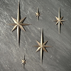 May include: A collection of gold-colored starburst wall decor in various sizes. The stars have multiple points and are arranged on a dark gray textured surface. The starbursts are a decorative accent.