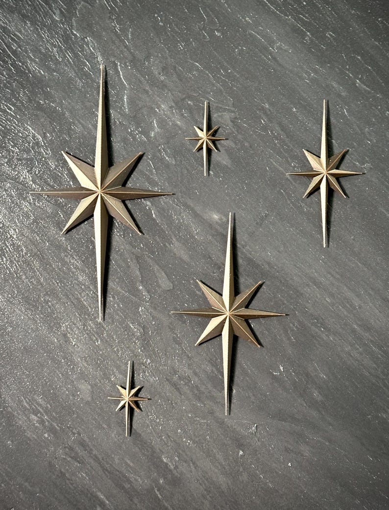 May include: A collection of gold-colored starburst wall decor in various sizes. The stars have multiple points and are arranged on a dark gray textured surface. The starbursts are a decorative accent.