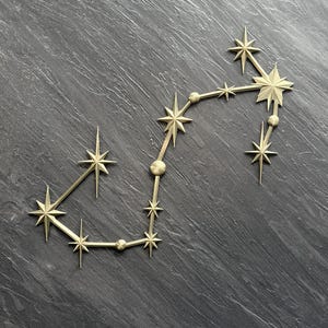 May include: A gold-colored metal wall decoration depicting the Scorpius constellation. The piece features a series of multi-pointed star shapes and small circular spheres connected by thin straight lines, forming the distinct celestial pattern.
