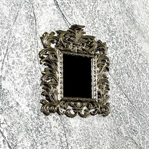 May include: A small, ornate mirror with a rectangular black reflective surface. The frame is a detailed, antique gold color with intricate scrollwork and floral designs. The mirror is a decorative accent piece.