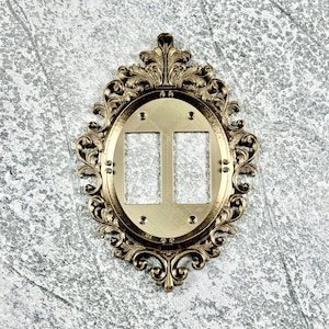 May include: A gold-colored double light switch plate cover featuring an ornate, oval-shaped frame with intricate scrollwork and acanthus leaf detailing. The central plate has two rectangular cutouts for switches and four small screw holes.
