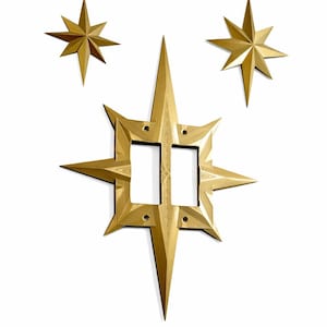 May include: Three gold-coloured star-shaped decorative pieces against a white background. The largest star has a square centre with two rectangular openings. Two smaller stars are positioned above the larger one.