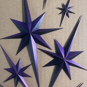 May include: An assortment of purple paper star ornaments in various sizes. The stars have sharp points and are arranged on a neutral background. The text "Packaged with Care" is at the bottom.